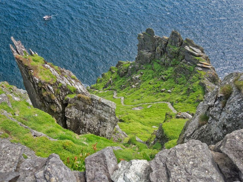 A high-angle view of a steep, grassy cliff with a winding stone path leading down towards a deep blue ocean with a boat.
