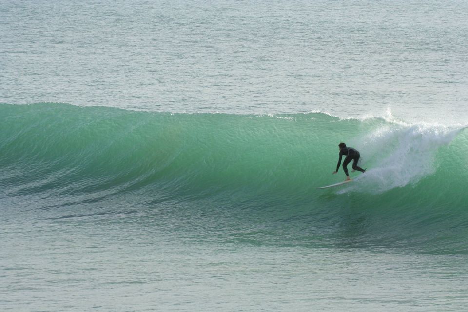 A surfer in a black wetsuit rides along the face of a large, green wave in the open ocean.