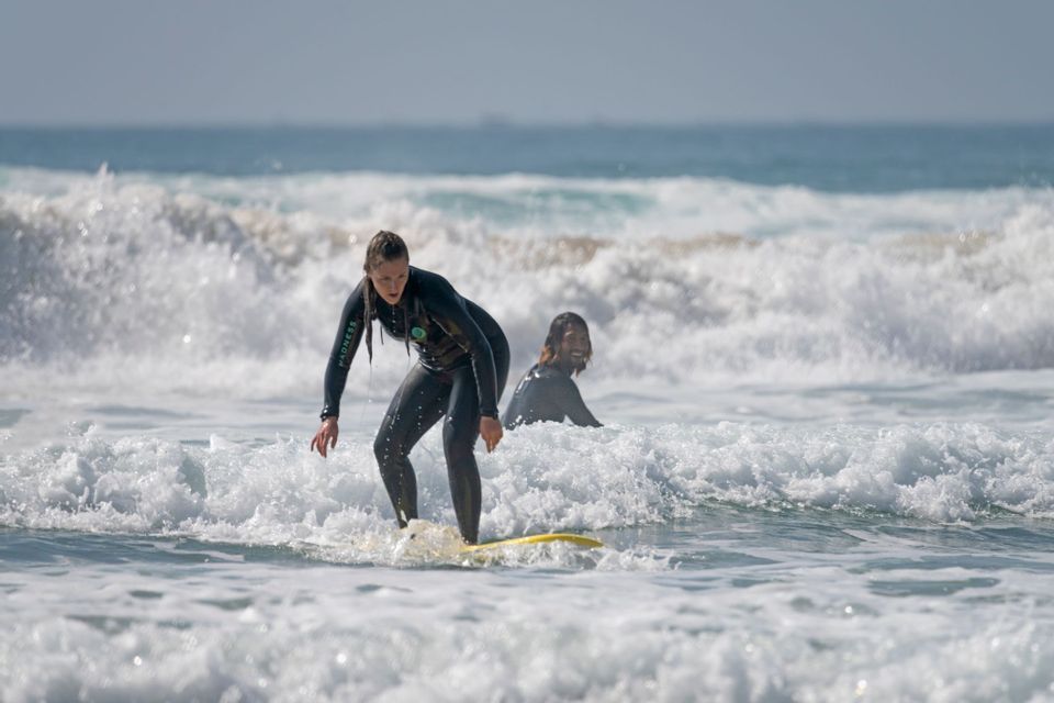 A woman in a wetsuit on a WeRoad group trip surfs on a yellow board, with another person smiling in the background waves.