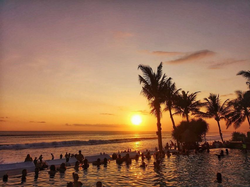 A WeRoad group trip of people in an infinity pool watching the sunset over the ocean with silhouetted palm trees.