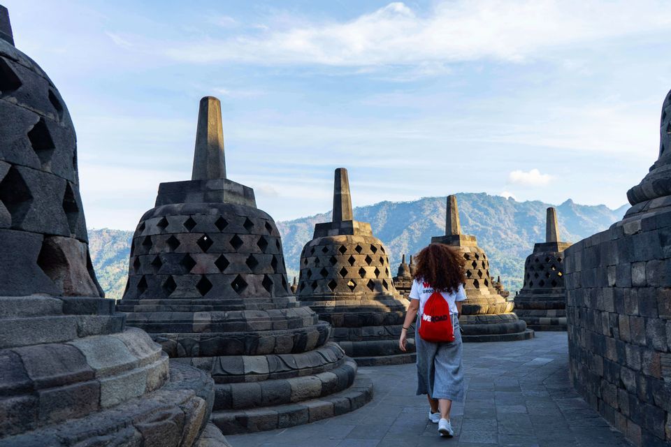 Eine Frau mit lockigem Haar und einem roten Rucksack geht auf einem Steinpfad zwischen großen, glockenförmigen Stupas, mit Bergen in der Ferne.