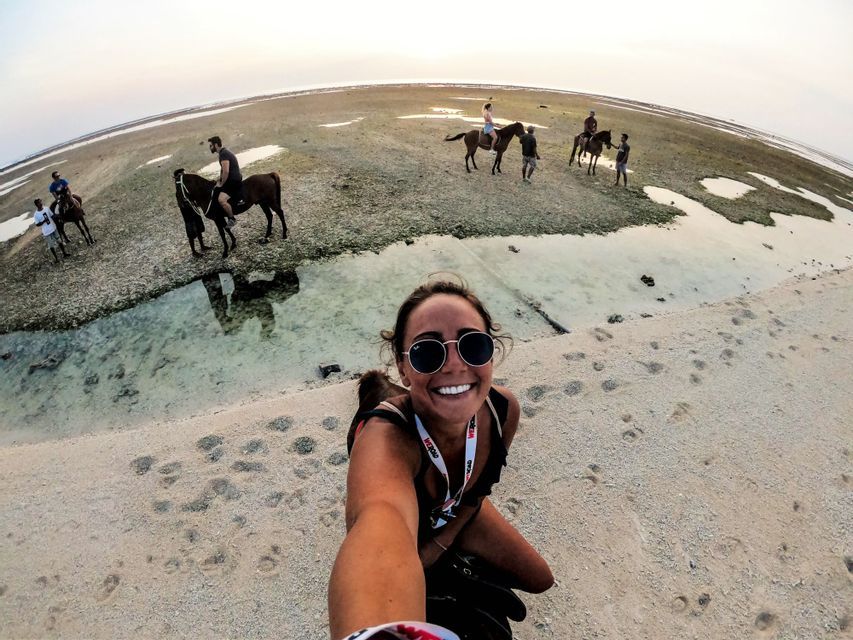 Une femme souriante prend un selfie sur une plage de sable, tandis qu'en arrière-plan, son groupe WeRoad fait une balade à cheval à marée basse.