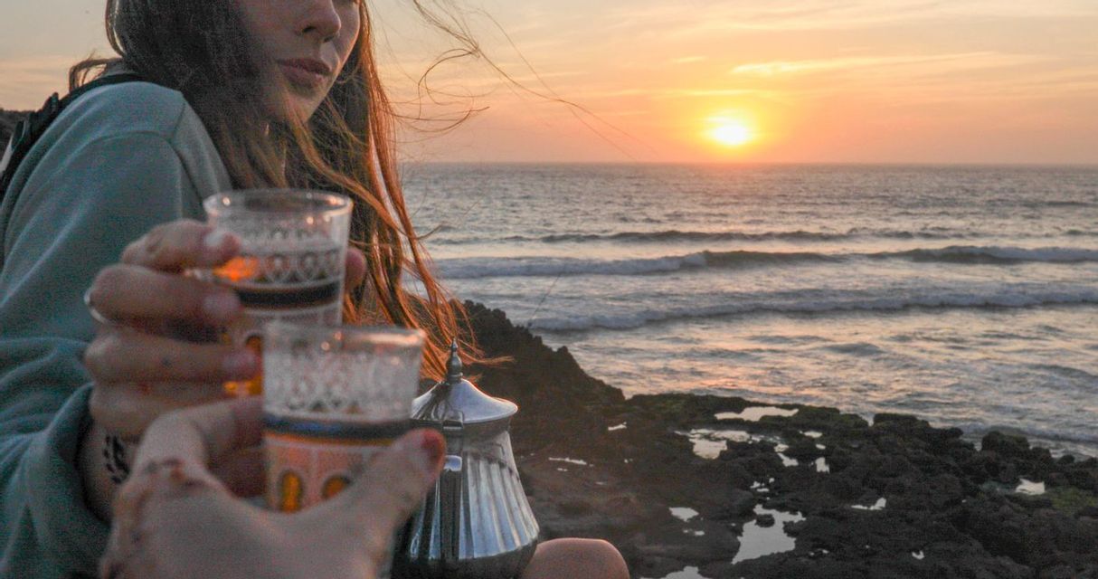 Two people toast with traditional tea glasses on a rocky coast as the sun sets over the ocean.