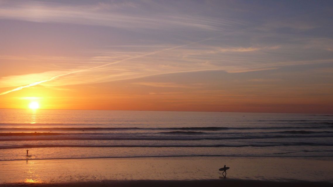 A silhouetted surfer carrying a surfboard walks along the shoreline during an orange sunset over the ocean.