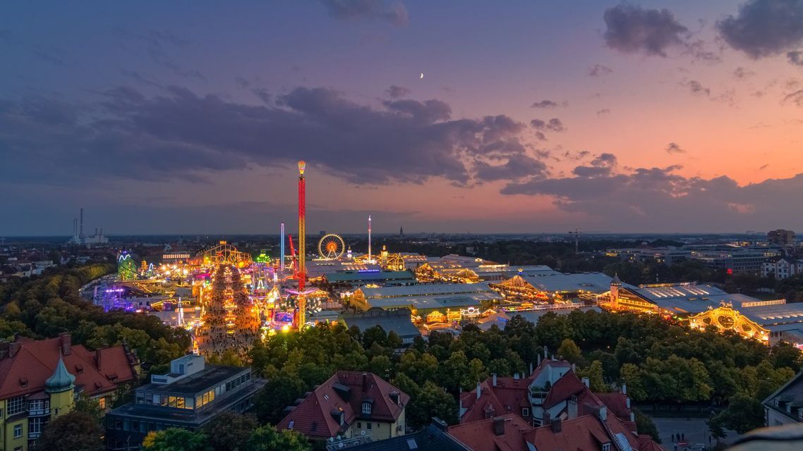 Vista aérea de un extenso y brillantemente iluminado parque de atracciones con juegos y atracciones al anochecer, con la silueta de la ciudad de fondo.