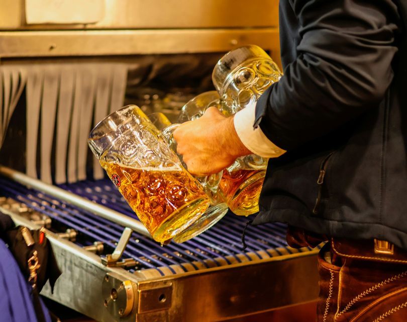 Una persona vestida con lederhosen y una chaqueta oscura sostiene varias jarras de cerveza grandes de cristal sobre una rejilla de lavado industrial.