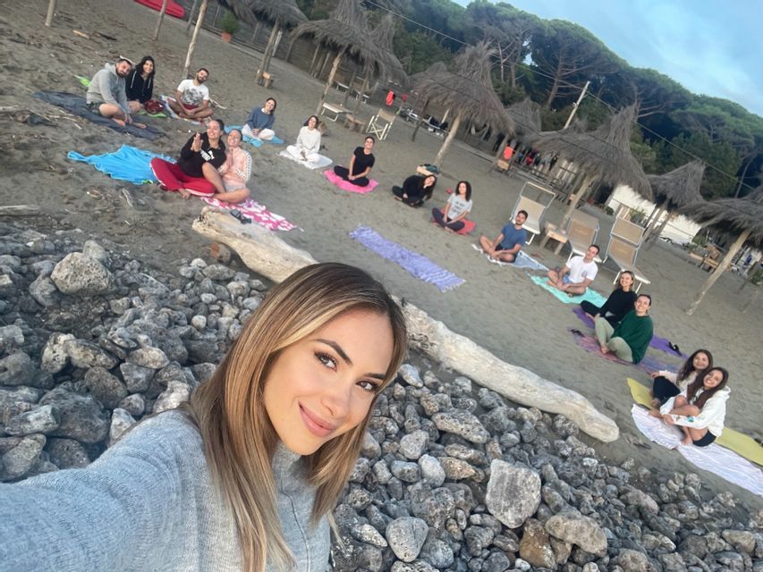 Una donna con un maglione grigio scatta un selfie su una spiaggia rocciosa mentre un gruppo di viaggio WeRoad è seduto su tappetini per una sessione di yoga.