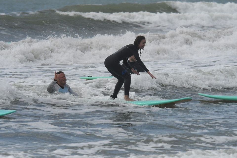 Una donna di un viaggio di gruppo WeRoad pratica surf, bilanciandosi sulla sua tavola mentre un uomo sorride dall'acqua increspata.