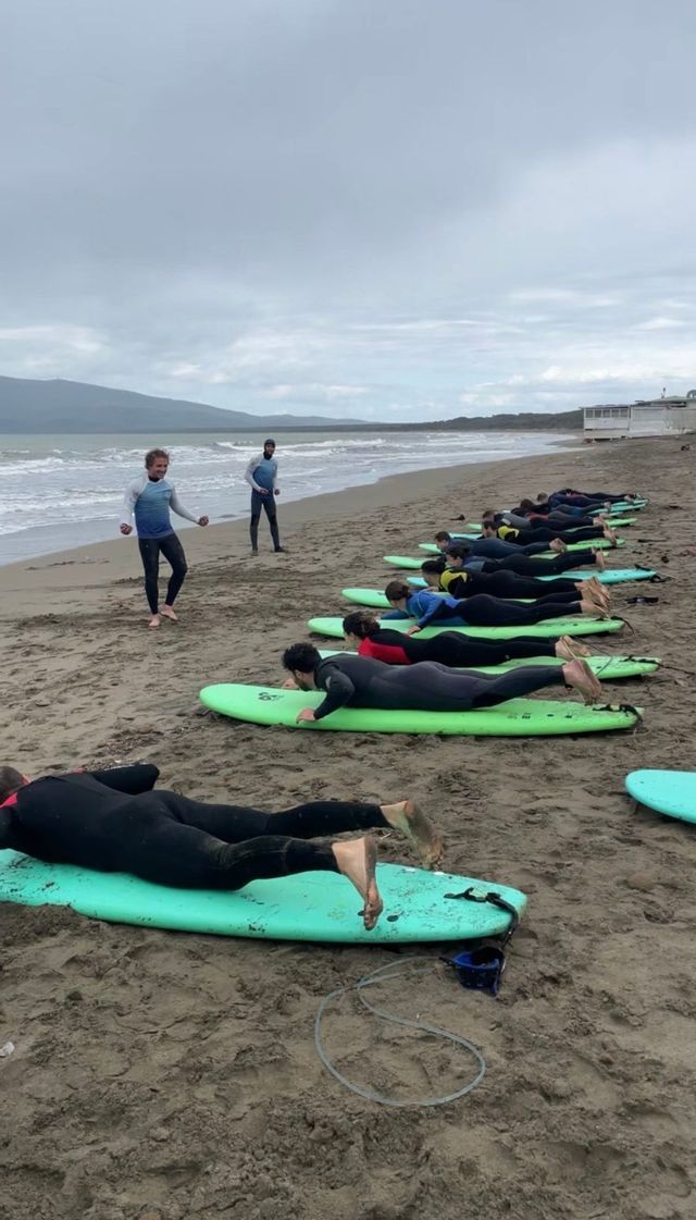 Un gruppo WeRoad prende una lezione di surf, sdraiato sulle tavole sulla sabbia mentre un istruttore li guida in riva al mare.