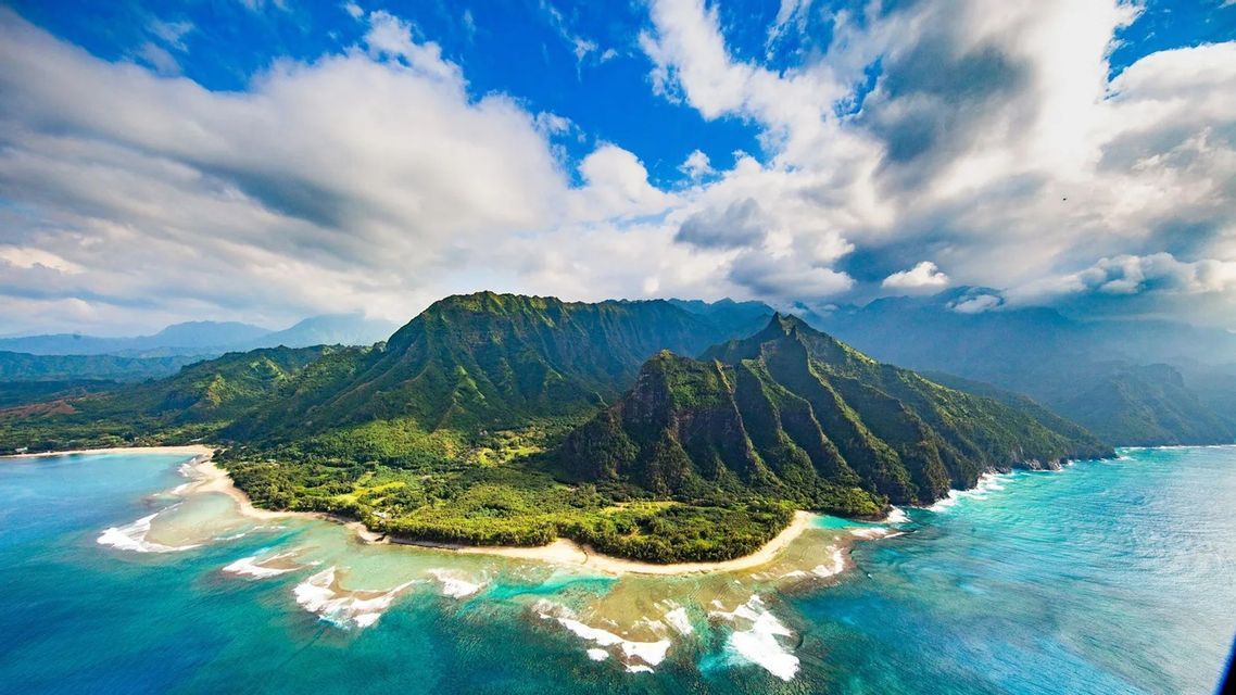 Veduta aerea di una costa montuosa e lussureggiante che incontra un oceano turchese, con spiagge sabbiose sotto un cielo azzurro nuvoloso.