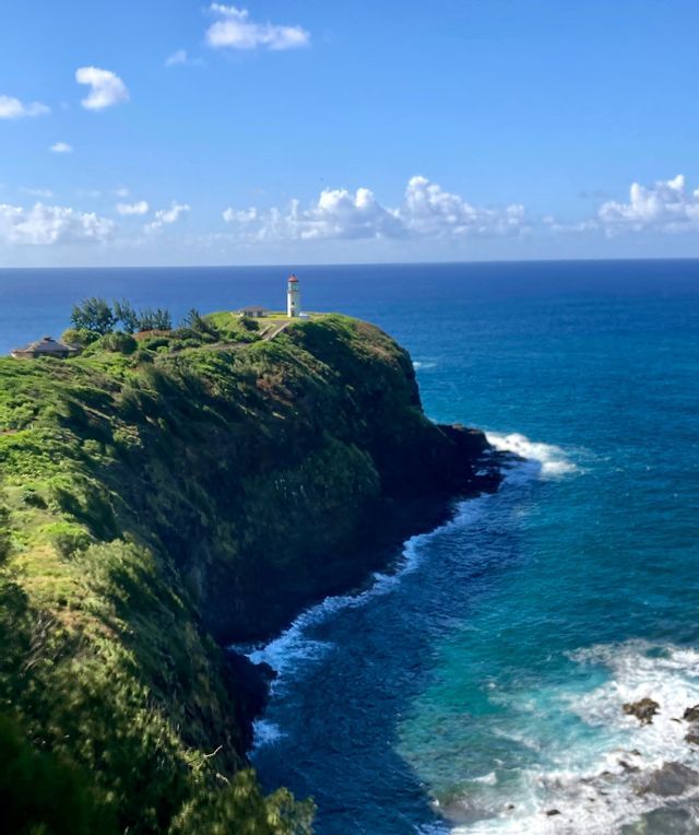Un faro bianco con la cima rossa si erge su una scogliera verde e lussureggiante che domina un vasto oceano blu sotto un cielo parzialmente nuvoloso.