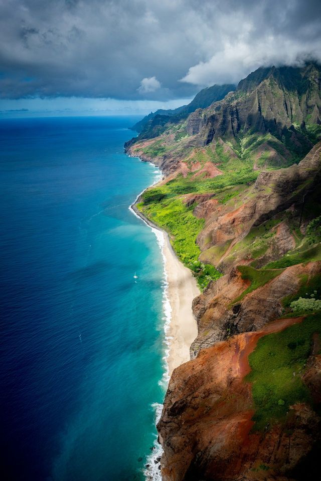 Vista aerea di una costa remota con ripide scogliere verdi, una spiaggia di sabbia bianca e acqua turchese sotto un cielo nuvoloso.