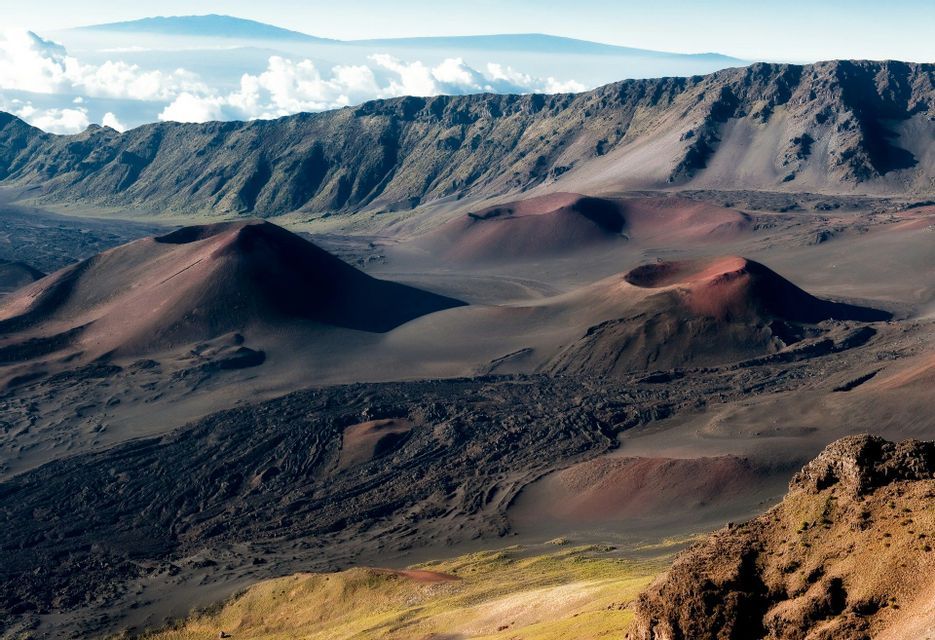 Un vasto paesaggio vulcanico caratterizzato da coni di scorie rossastre e colate di lava scure all'interno di una grande caldera sotto un cielo limpido.