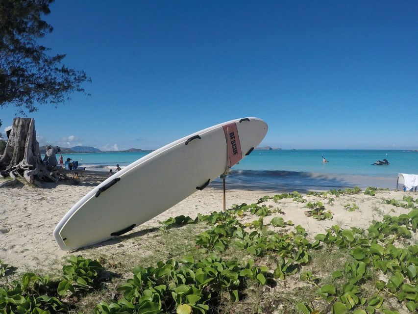 Una tavola da surf bianca da salvataggio si erge dritta su una spiaggia sabbiosa con acqua turchese sotto un cielo azzurro e limpido.