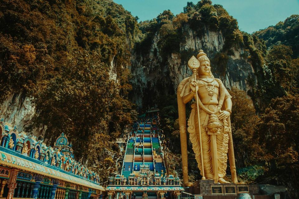 Eine große goldene Statue einer Gottheit steht neben einer langen, farbenfrohen Treppe, die einen bewaldeten Felsen hinaufführt.