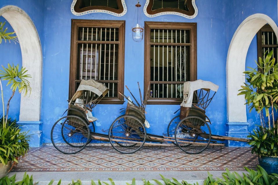 Zwei traditionelle Rikschas parkten auf einer gefliesten Veranda vor einer leuchtend blauen Wand mit großen Holzfenstern.