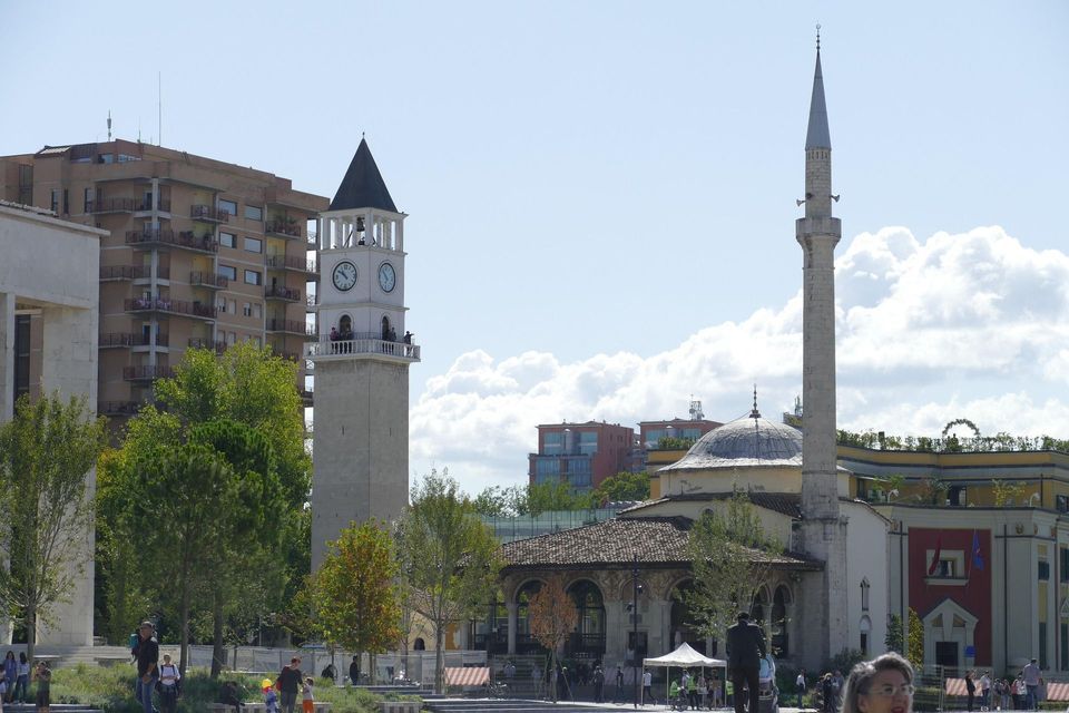 Una storica torre dell'orologio in pietra e una moschea con minareto si ergono in una piazza cittadina circondata da edifici moderni.