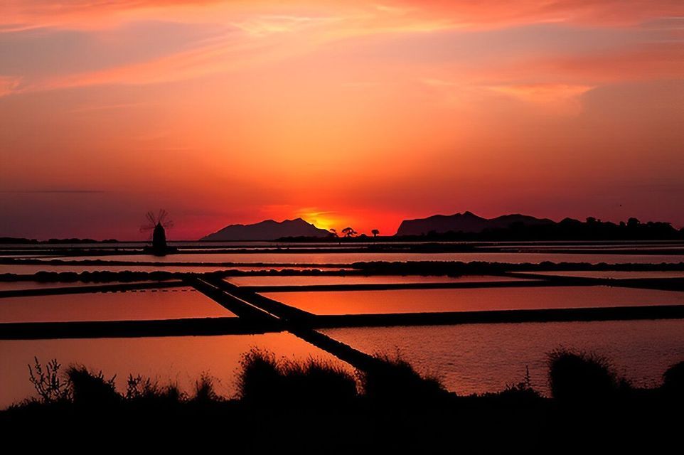 Un molino de viento silueteado se alza entre las salinas, reflejando un vibrante atardecer naranja sobre montañas distantes.