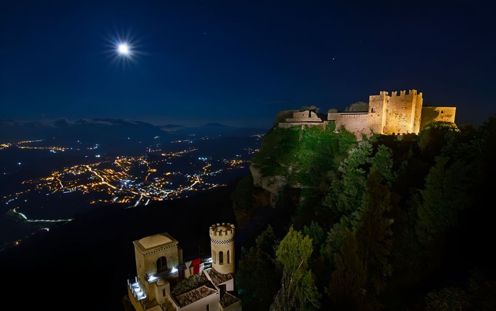 Un castillo iluminado en un acantilado domina un valle de luces de la ciudad por la noche bajo una luna brillante.