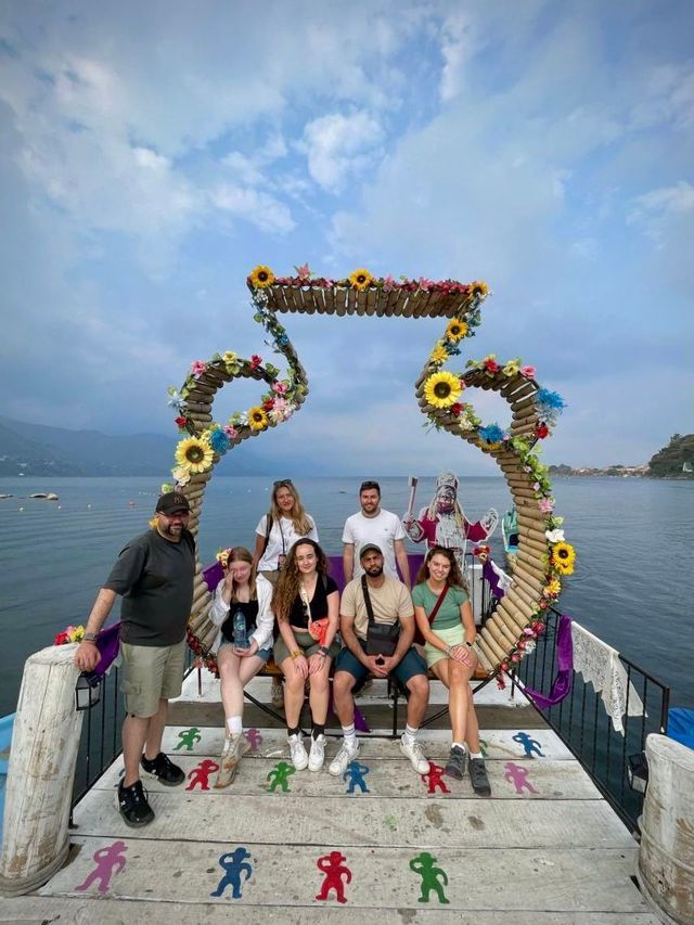 A WeRoad group trip poses for a photo on a wooden pier by a lake, in front of a large, flower-adorned sculpture.