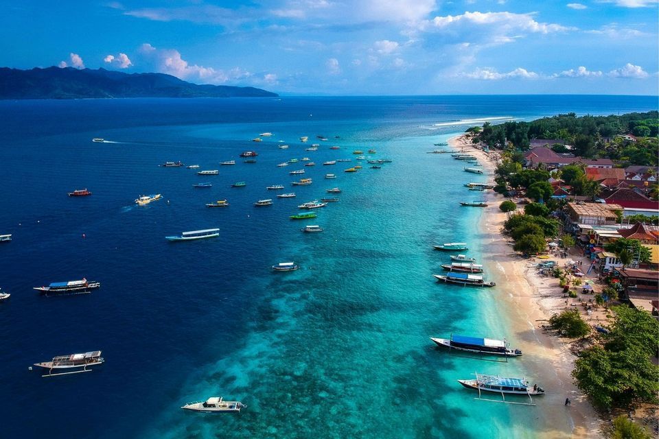 Luftaufnahme vieler Boote in klarem, türkisfarbenem Wasser an einem Sandstrand mit Gebäuden und Bäumen.