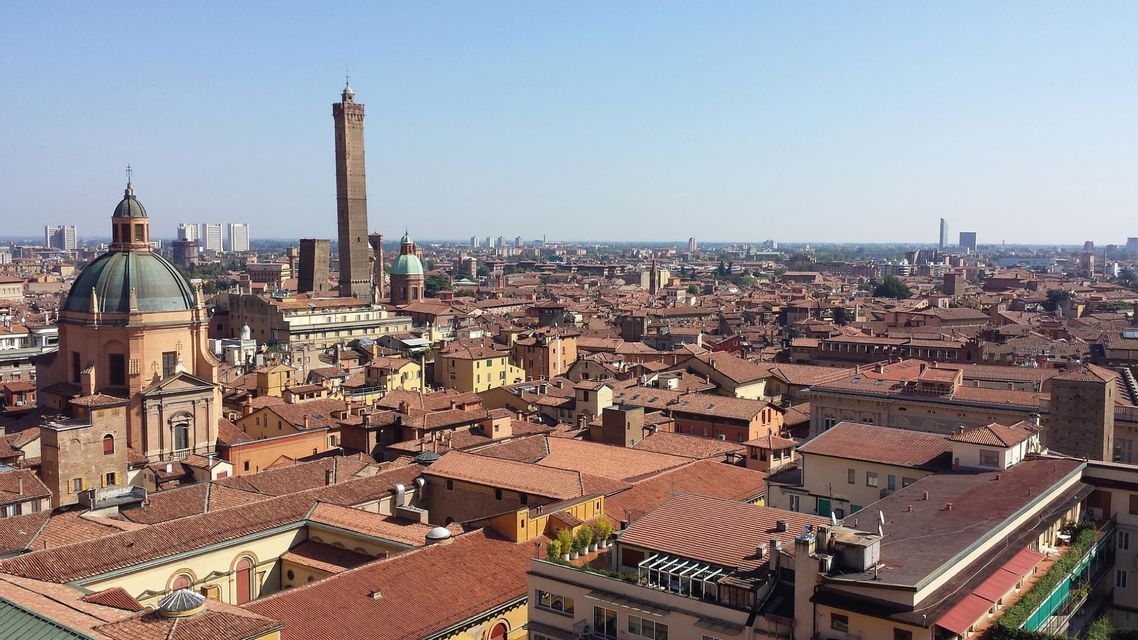 Una vista panoramica di una città con tetti di tegole rosse, un'alta torre di mattoni e una cupola prominente sotto un cielo sereno.