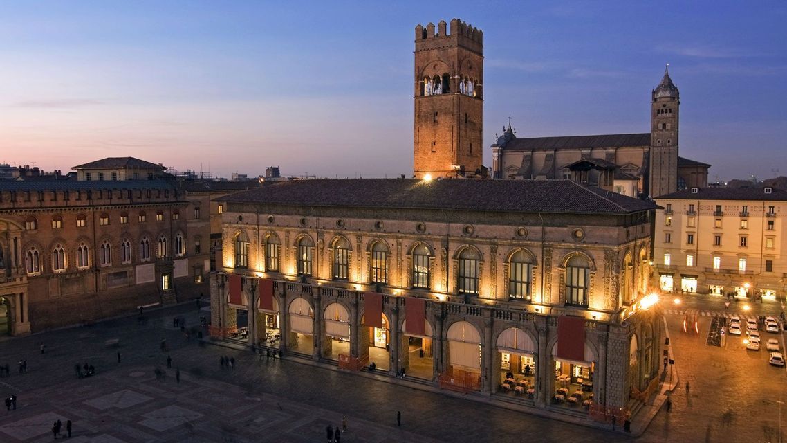 Una vista dall'alto di una piazza storica cittadina al crepuscolo, con edifici illuminati e un'imponente torre in mattoni