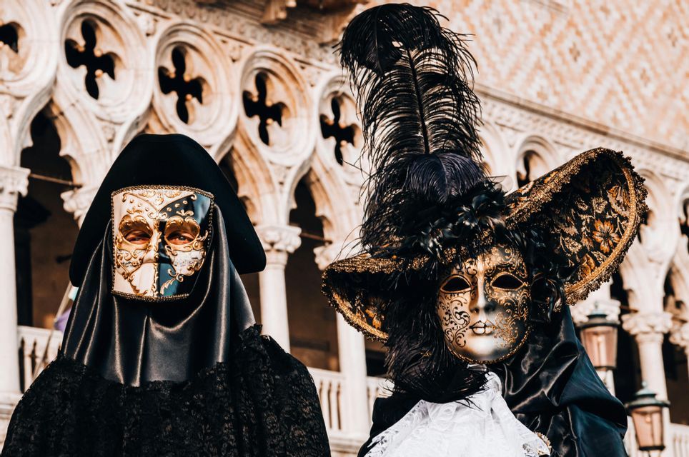 Deux personnes en costumes de carnaval vénitiens ornés, avec des masques élaborés noirs et dorés, posent devant un bâtiment aux arches blanches.