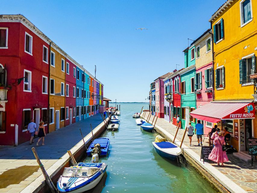 Un canal ensoleillé, avec ses bateaux amarrés, serpente entre deux rangées de bâtiments aux couleurs vives, tandis que des gens flânent le long des quais.
