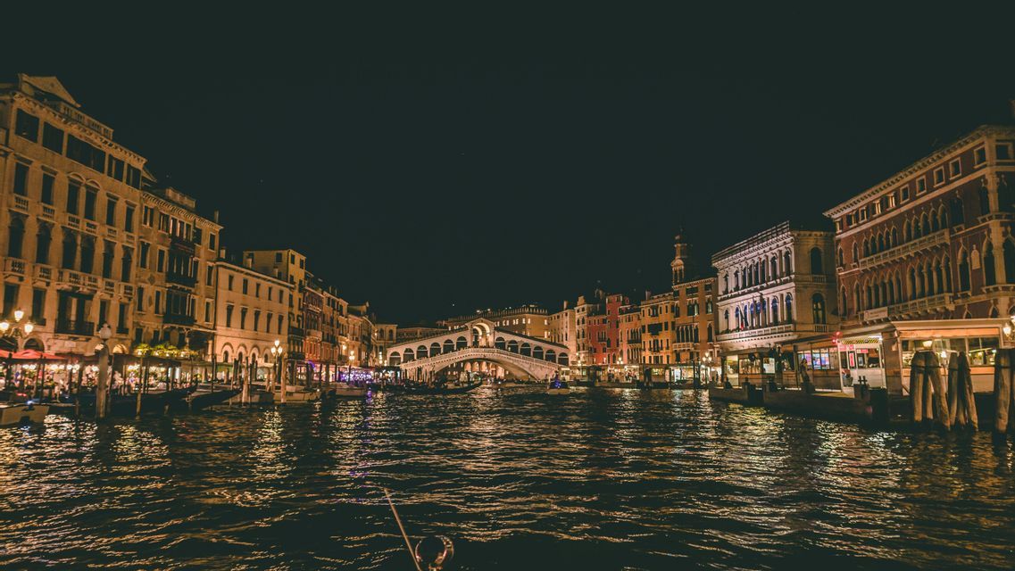 Un large canal la nuit avec des bâtiments historiques et un grand pont arqué, le tout illuminé et se reflétant dans l'eau.