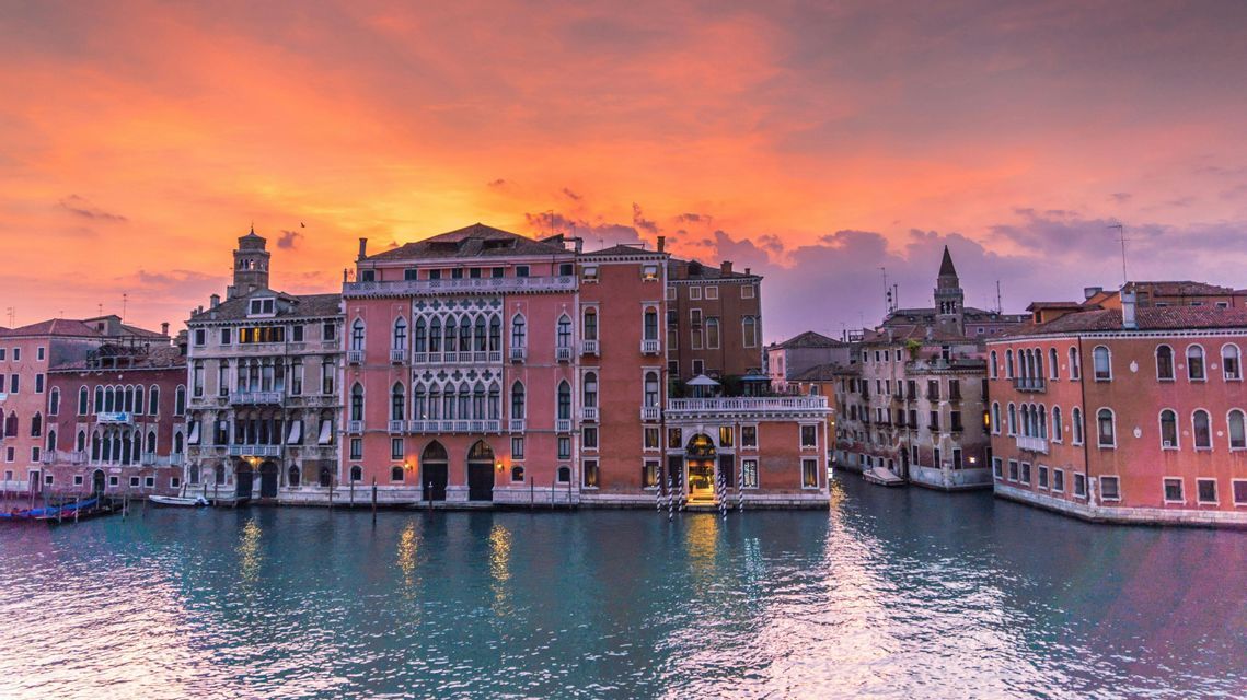 Des bâtiments historiques à l'architecture ornée bordent un canal sous un ciel de coucher de soleil orange et violet éclatant.
