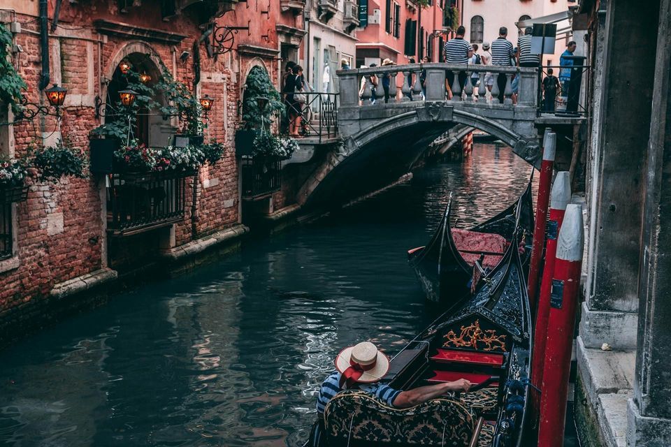 A gondolier in a straw hat steers a gondola down a narrow canal between brick buildings, with a stone bridge in the background.