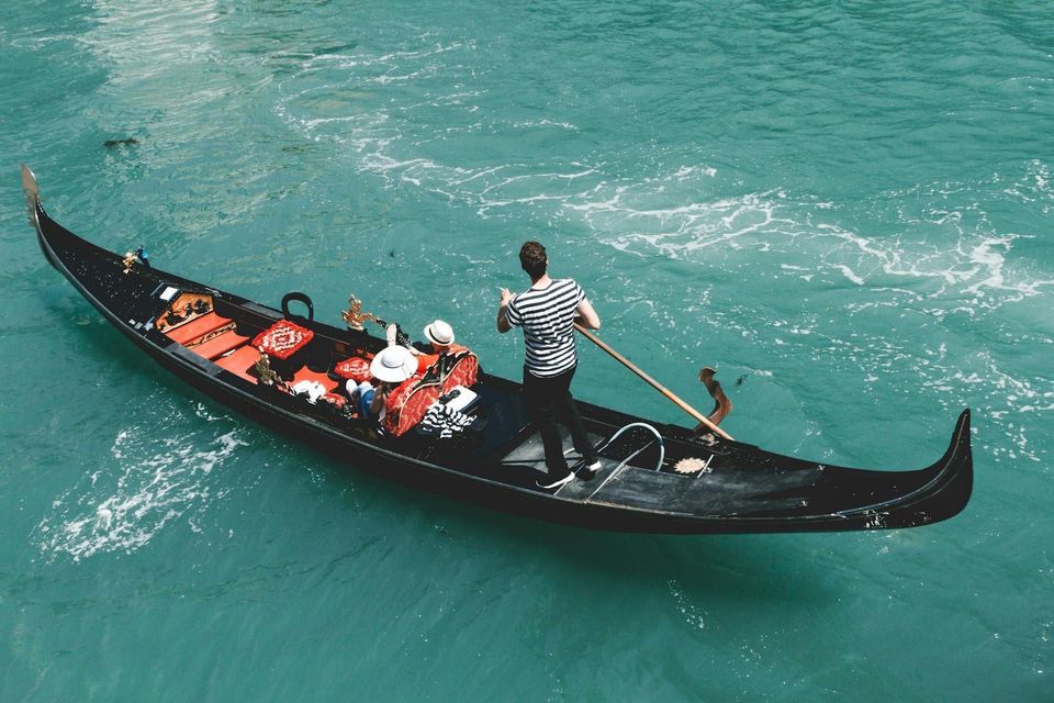 A gondolier in a striped shirt rows a gondola with two passengers from a WeRoad group trip across turquoise water.