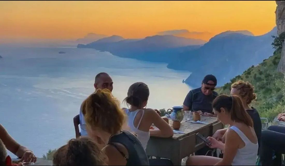 Un viaje en grupo de WeRoad se reúne en una mesa de madera con vistas a una costa montañosa y al mar al atardecer.