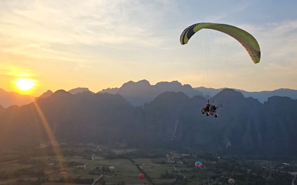 Un parapendio a motore vola su una valle montuosa all'alba, con diverse mongolfiere visibili sotto.