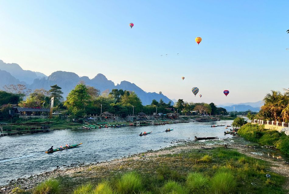 Mongolfiere fluttuano in un cielo limpido sopra un ampio fiume con barche a coda lunga, incorniciato da alberi lussureggianti e montagne lontane.
