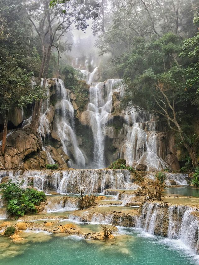 Una cascata a più livelli precipita lungo sporgenze rocciose in pozze turchesi, circondata da una foresta lussureggiante e nebbiosa.