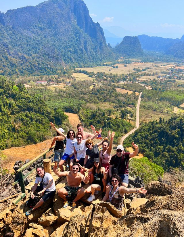 Un gruppo WeRoad posa insieme su un punto panoramico roccioso che si affaccia su una vasta valle verde circondata da montagne.