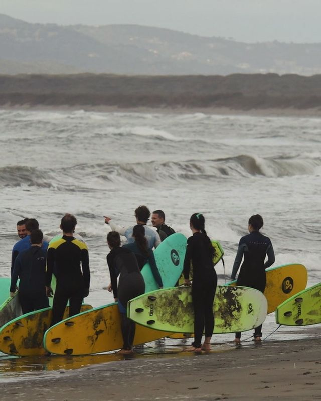 Un viaggio di gruppo WeRoad con persone in muta e tavole da surf su una spiaggia, mentre osservano le onde agitate.