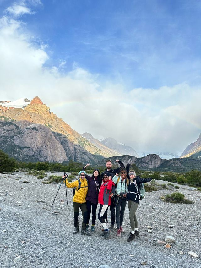 A WeRoad group trip in hiking gear poses in a rocky mountain valley with a faint rainbow in the background.