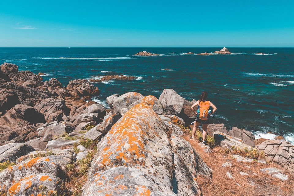 Una mujer con una mochila de pie en una costa rocosa, contemplando el océano azul profundo bajo un cielo despejado.