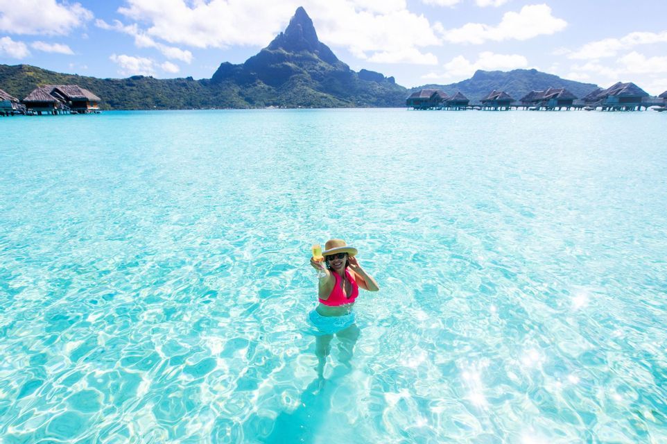 Eine Frau mit Hut steht in klarem türkisfarbenem Wasser, mit Überwasser-Bungalows und einem Berg im Hintergrund.