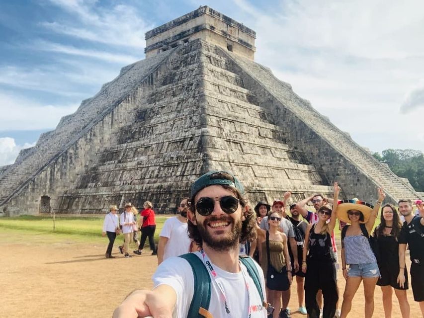 A man takes a selfie with his WeRoad group trip as they pose in front of a large stone pyramid under a partly cloudy sky.