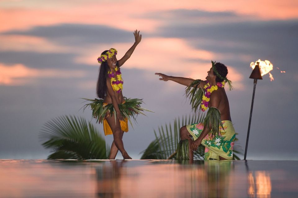 Ein Mann und eine Frau in traditioneller polynesischer Kleidung tanzen bei Sonnenuntergang an einem Infinity-Pool mit einer beleuchteten Tiki-Fackel.