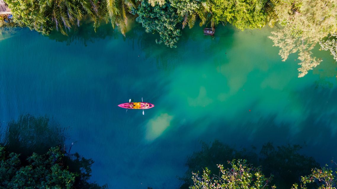 Luftaufnahme von zwei Personen, die ein rosa Kajak auf einem türkisfarbenen und blauen Fluss paddeln, umgeben von üppig grünen Bäumen.