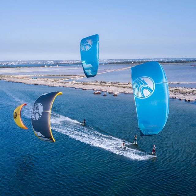 Un grupo de WeRoad practica kitesurf en aguas azules con cometas grandes y coloridas, visto desde arriba.