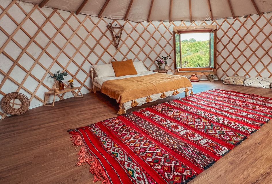 L'interno di una yurta con un letto fatto, un grande tappeto rosso a motivi su un pavimento di legno e una finestra che si affaccia su colline verdi.
