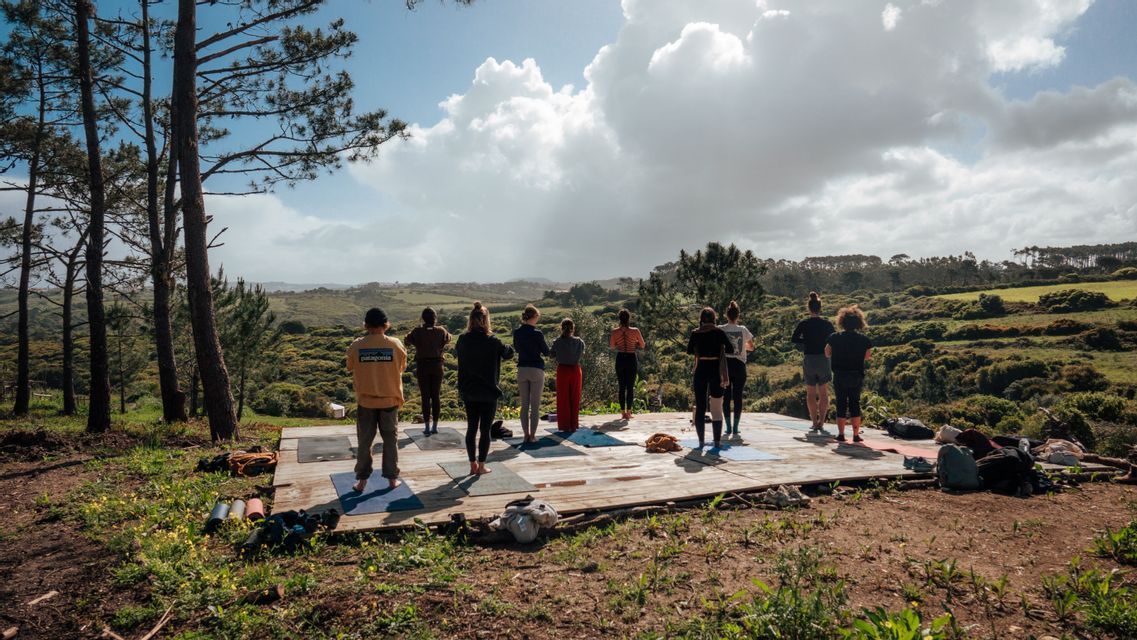 Un gruppo WeRoad pratica yoga su una piattaforma di legno all'aperto con vista su un paesaggio verde e collinare sotto un cielo nuvoloso.