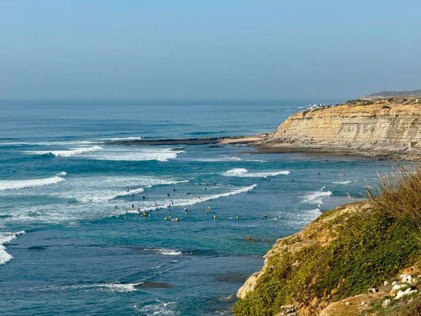 Surfisti sulle loro tavole sono sparsi su un oceano turchese con onde bianche che si infrangono vicino a un'alta scogliera sabbiosa.