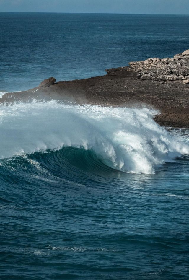 Una grande onda blu si increspa e si infrange con schiuma bianca contro una costa rocciosa.